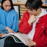 Two students reading and discussing books in a library setting for their study session.