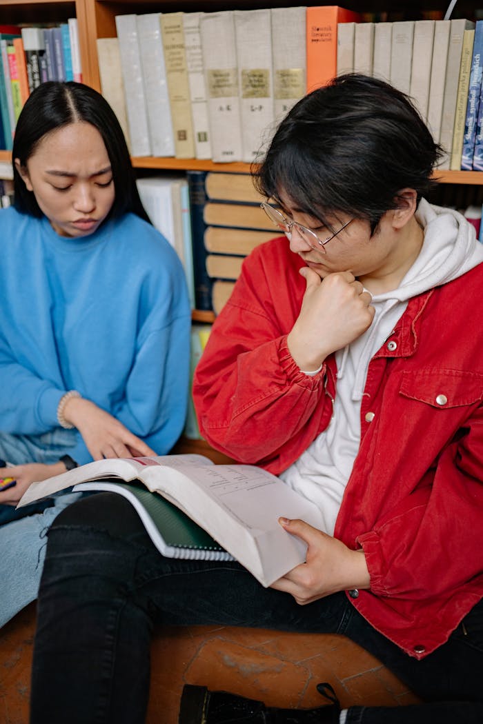 Two students reading and discussing books in a library setting for their study session.